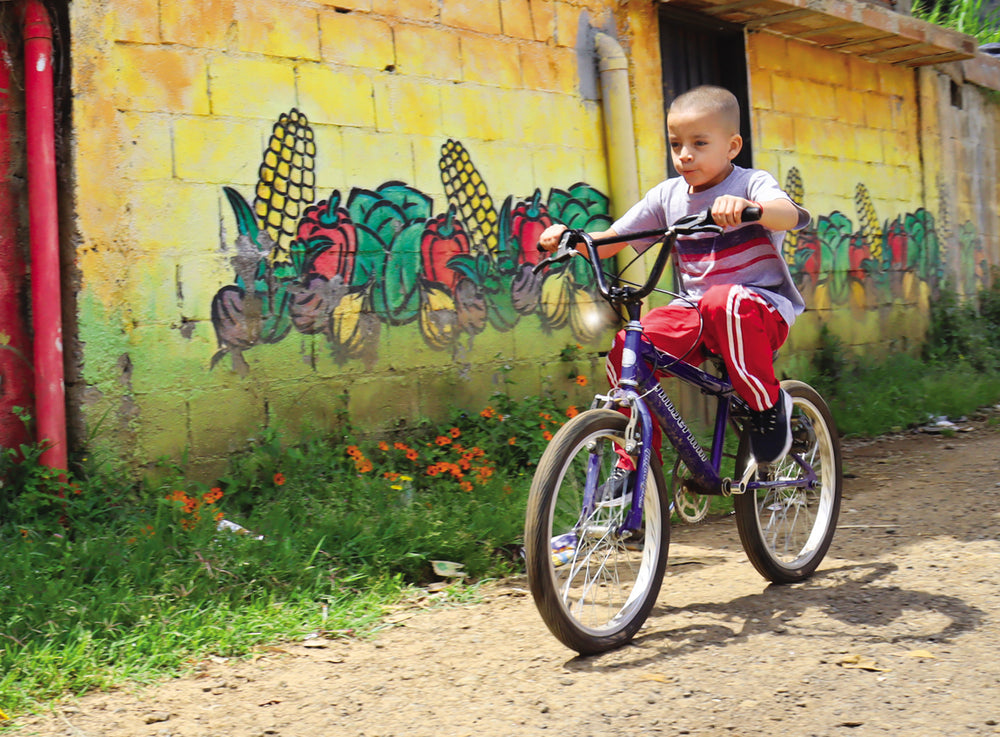 Un jeune garçon fait du vélo dans une allée, une peinture murale représentant des légumes se trouvant derrière lui.