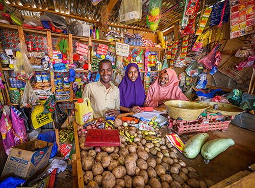 Un homme et deux femmes sourient. Ils se tiennent dans un magasin qu’ils gèrent, rempli de divers biens et fruits et légumes.