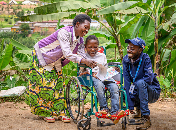 Une femme et un homme sourient et jouent avec enfant en fauteuil roulant à l’extérieur.