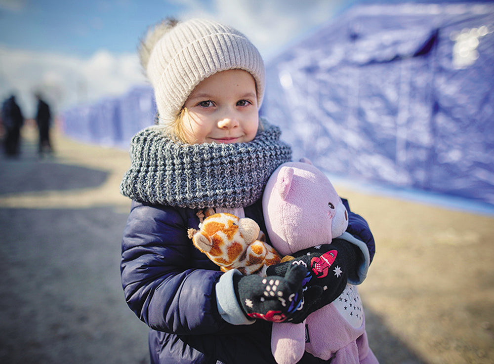 Une jeune fille habillée avec des vêtements chauds se tient devant des tentes. Elle serre des animaux en peluche contre elle.