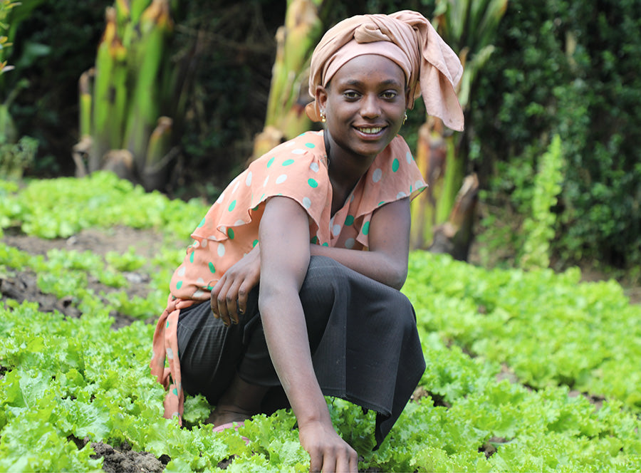 Une femme sourit, accroupie devant un espace entouré de verdure.